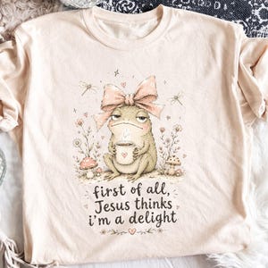 May include: A light beige t-shirt featuring a graphic of a frog wearing a large pink bow, holding a steaming mug with a heart icon. The design includes mushrooms, dragonflies, and the text: first of all, Jesus thinks i'm a delight.