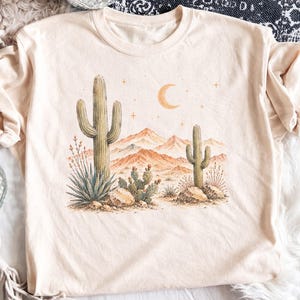 以下が含まれることがあります： 砂漠の風景が描かれたライトベージュのTシャツ。プリントには、2本の背の高いサワロサボテン、小さめのウチワサボテン、岩、遠くの山々が、三日月といくつかの小さな四芒星の下に描かれています。