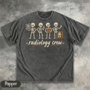 Pode incluir: Uma t-shirt cinzenta escura de manga curta com um gráfico de quatro esqueletos. Os esqueletos seguram um raio-x, fazem o sinal de paz, seguram uma abóbora e fazem um sinal de positivo. O texto "radiology crew" está impresso abaixo em tom claro.
