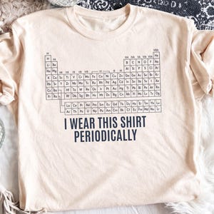 Peut inclure: Un t-shirt à manches courtes beige clair avec un graphique noir du tableau périodique des éléments imprimé sur le devant. Sous le tableau, le texte I WEAR THIS SHIRT PERIODICALLY est imprimé dans une police sans empattement bleu foncé.