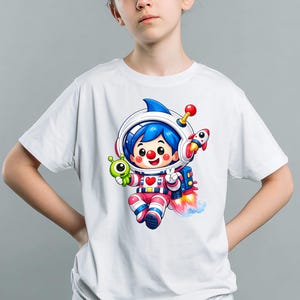 May include: White t-shirt featuring a cartoon astronaut character with blue hair, a space helmet, and a rocket. The astronaut is holding a green alien and a rocket ship. The design includes red, pink, and blue accents.