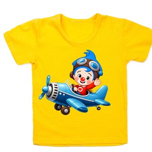 Puede incluir: Camiseta amarilla de manga corta con una ilustraci&oacute;n de dibujos animados de un personaje en un avi&oacute;n azul. El personaje tiene pelo azul, gafas y una camisa roja. El avi&oacute;n es azul con una h&eacute;lice.