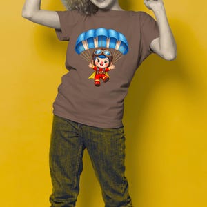 Puede incluir: Camiseta marr&oacute;n con una ilustraci&oacute;n de dibujos animados de un personaje con un paraca&iacute;das azul. El personaje lleva un atuendo rojo y amarillo con gafas. El fondo es de color amarillo liso.