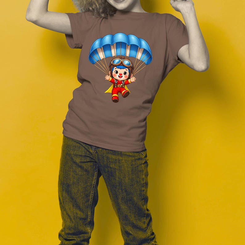 Puede incluir: Camiseta marr&oacute;n con una ilustraci&oacute;n de dibujos animados de un personaje con un paraca&iacute;das azul. El personaje lleva un atuendo rojo y amarillo con gafas. El fondo es de color amarillo liso.