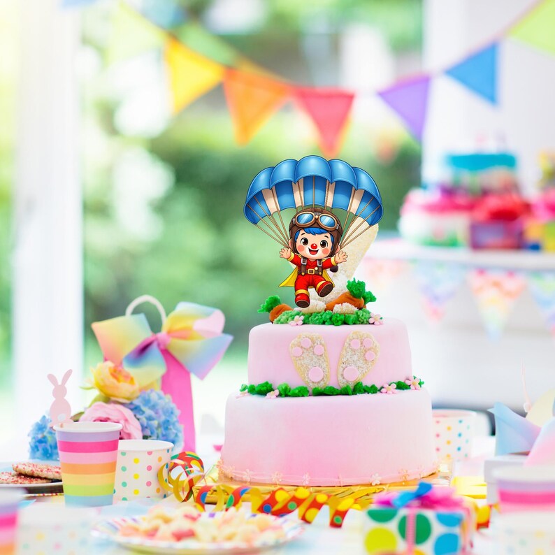 Puede incluir: Un pastel rosa de dos niveles decorado con una figura de dibujos animados con un paraca&iacute;das azul. El pastel est&aacute; decorado con glaseado verde y peque&ntilde;as flores rosas. Decoraciones coloridas de fiesta en el fondo.