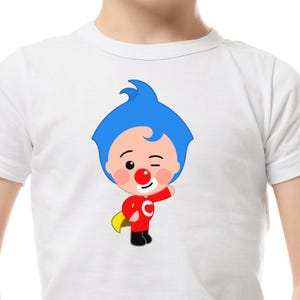 May include: White t-shirt featuring a cartoon character with blue hair, a red nose, and a red outfit with a yellow cape. The character is winking and has a smiling expression. The shirt is made of a soft, comfortable material.