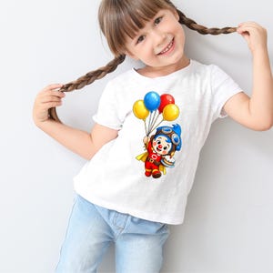 Puede incluir: Camiseta blanca con un dise&ntilde;o de payaso de dibujos animados sosteniendo globos y un pastel. El payaso lleva un casco de aviador azul, un traje rojo y una capa amarilla. Los globos son amarillos, azules y rojos. La camiseta la lleva un ni&ntilde;o peque&ntilde;o.