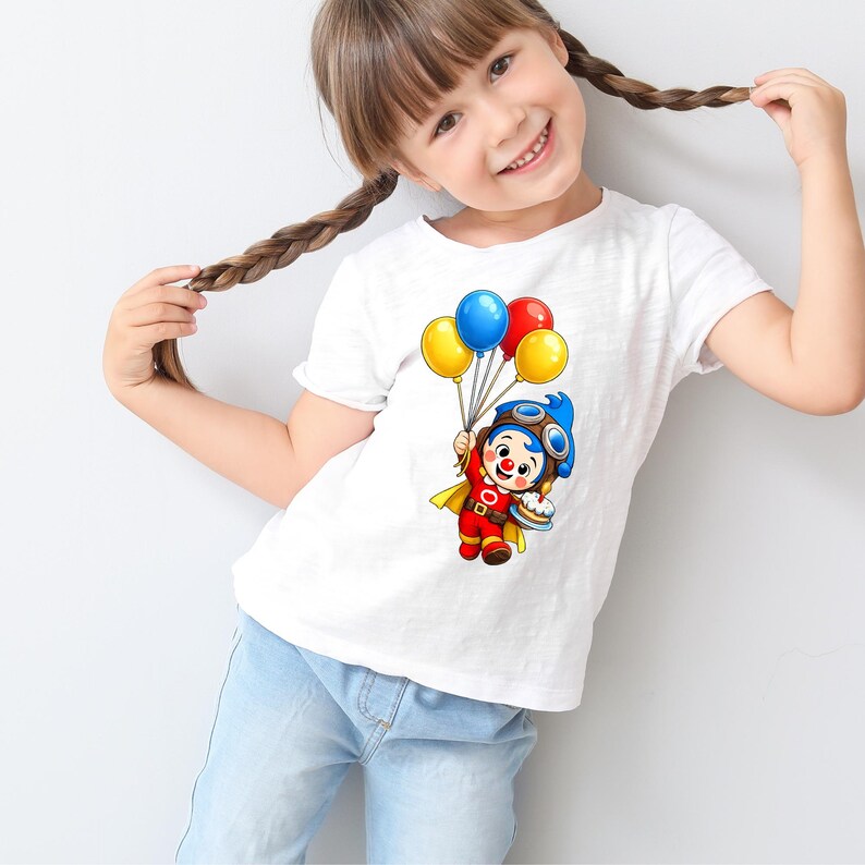 Puede incluir: Camiseta blanca con un dise&ntilde;o de payaso de dibujos animados sosteniendo globos y un pastel. El payaso lleva un casco de aviador azul, un traje rojo y una capa amarilla. Los globos son amarillos, azules y rojos. La camiseta la lleva un ni&ntilde;o peque&ntilde;o.
