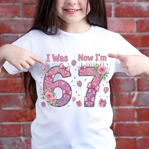 May include: White t-shirt with the text "I Was Now I'm" above the numbers 67. The numbers are decorated with pink glitter, bows, flowers, and birthday candles. Balloons and gift boxes are also featured.