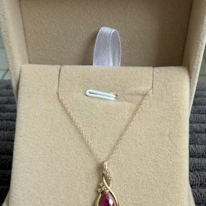 May include: A gold pendant necklace featuring a teardrop-shaped ruby gemstone. The pendant is set in a gold frame and embellished with small diamonds. The necklace is displayed in a beige jewellery box.