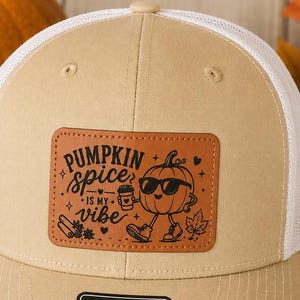 May include: A tan and white trucker hat featuring a rectangular leather patch with a black graphic of a pumpkin wearing sunglasses and holding a coffee cup. The text reads PUMPKIN spice IS MY vibe. A black sticker on the brim displays the text 112 The Original MD-LG.