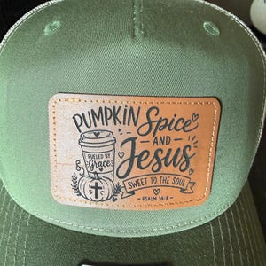 May include: An olive green baseball cap featuring a rectangular tan leather patch on the front. The patch displays a graphic of a coffee cup and pumpkin with the text Pumpkin Spice and Jesus, Fueled by Grace, Sweet to the Soul, Psalm 34:8.
