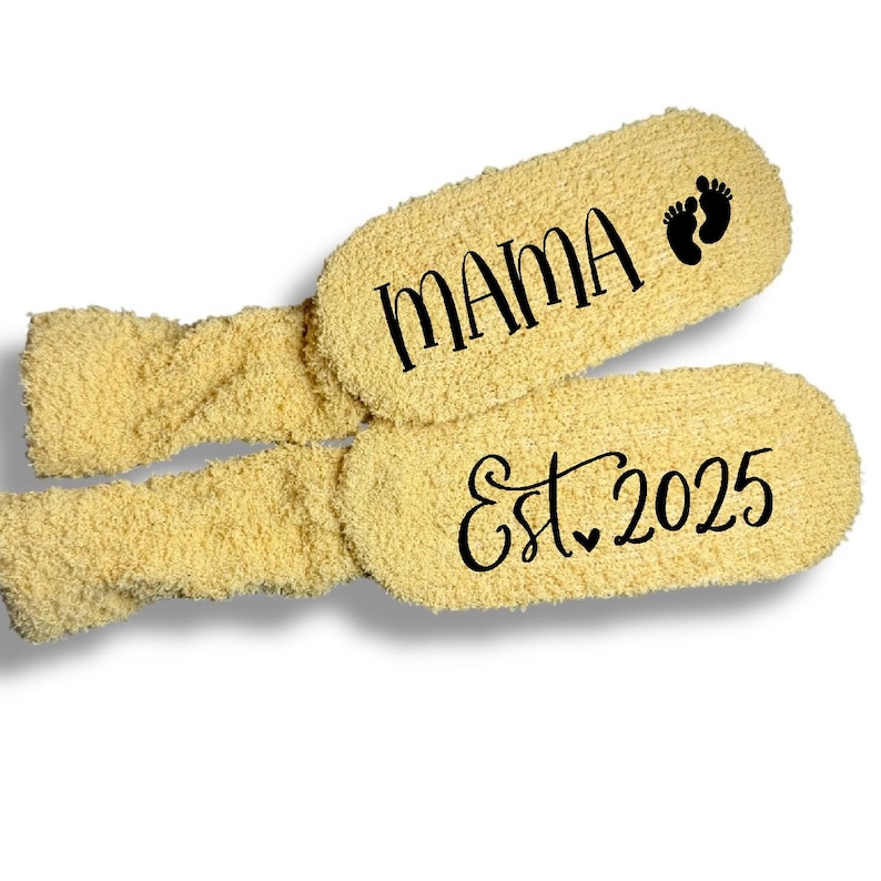 May include: A pair of fuzzy yellow socks featuring black text and graphics. One sock displays the word MAMA with a small footprint icon, while the other sock features the text Est. 2025 with a small heart symbol integrated into the lettering.
