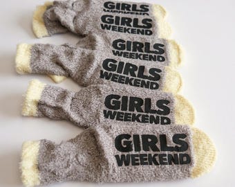 May include: Four pairs of fuzzy grey socks with cream-colored cuffs. Each sock features the text GIRLS WEEKEND printed in bold black lettering on the top. The socks have a soft, textured appearance and are arranged in a vertical stack on a plain surface.