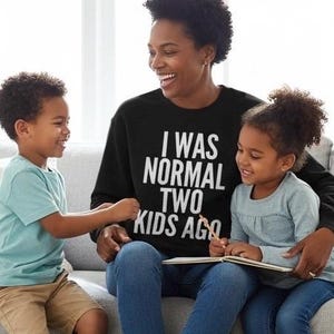 May include: A black sweatshirt with the text "I WAS NORMAL TWO KIDS AGO" in white, worn by a person sitting with two children. The children are looking at a book. One child is wearing a light blue t-shirt and khaki shorts.