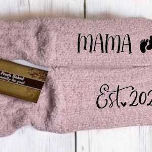 May include: A pair of soft, light pink socks with the word "MAMA" and baby footprints printed on one sock, and "Est. 2025" on the other. A small gift tag is placed on the socks.