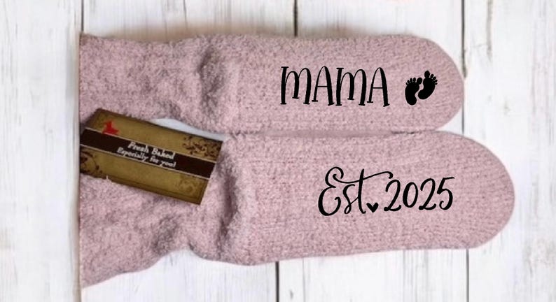 May include: A pair of soft, light pink socks with the word "MAMA" and baby footprints printed on one sock, and "Est. 2025" on the other. A small gift tag is placed on the socks.