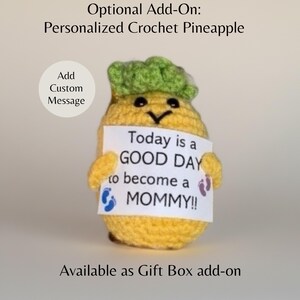 May include: A crocheted pineapple with a green top and yellow body holds a sign that reads "Today is a GOOD DAY to become a MOMMY!!" The sign has blue and purple footprints. The text "Optional Add-On: Personalized Crochet Pineapple" is above the pineapple.