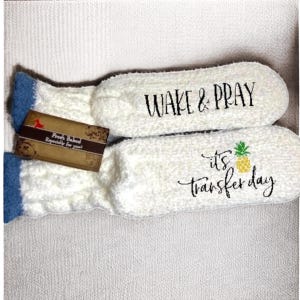 May include: A pair of white, fluffy socks with blue trim. One sock has the text "WAKE & PRAY" in black script. The other sock has a pineapple graphic and the text "it's transfer day". A small tag is attached.