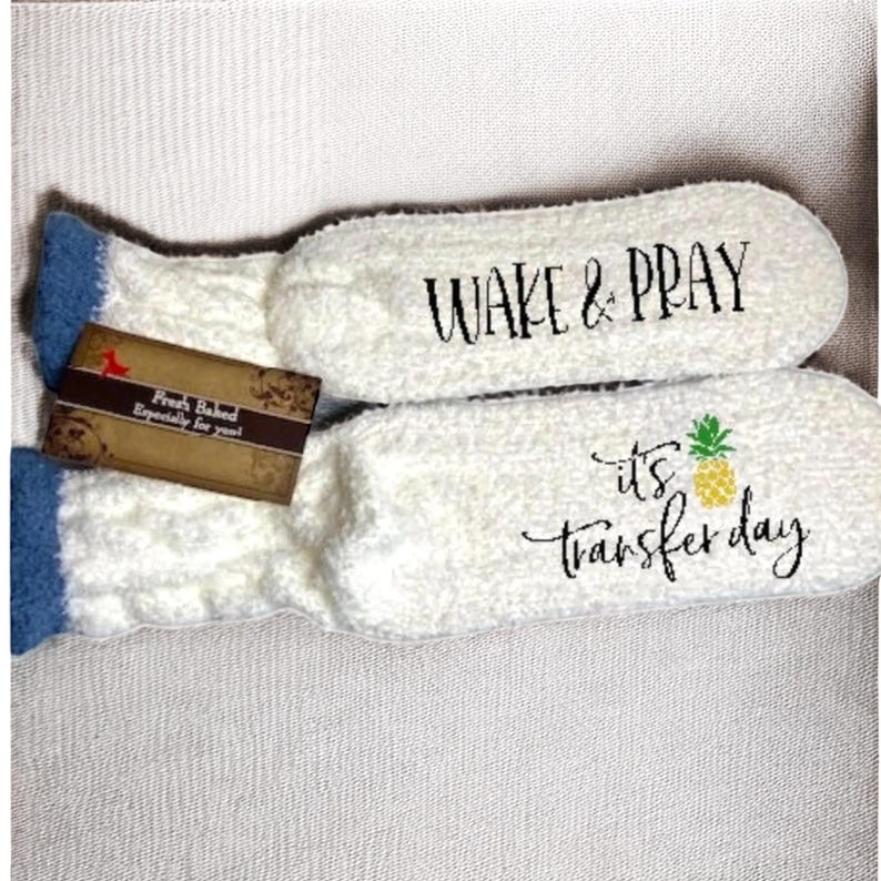 May include: A pair of white, fluffy socks with blue trim. One sock has the text "WAKE & PRAY" in black script. The other sock has a pineapple graphic and the text "it's transfer day". A small tag is attached.