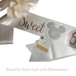 May include: A white satin sash featuring the rose gold text Sweet 16 with a rhinestone-encrusted mouse head icon positioned between the words. A small square card with a QR code and gold glitter accent is attached to the fabric surface.