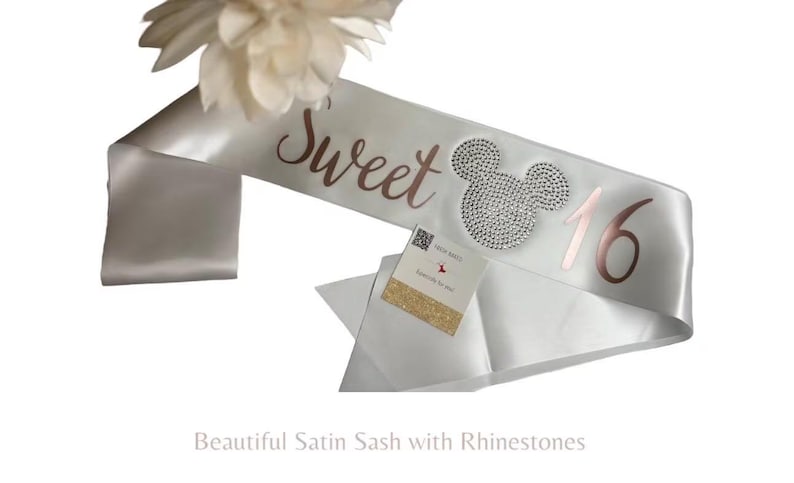 May include: A white satin sash featuring the rose gold text Sweet 16 with a rhinestone-encrusted mouse head icon positioned between the words. A small square card with a QR code and gold glitter accent is attached to the fabric surface.
