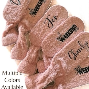 May include: Pink fuzzy slippers with names and "Girl's Weekend 2024" printed on them. The slippers are arranged in a fan shape. The text "Multiple Colors Available" is also visible.
