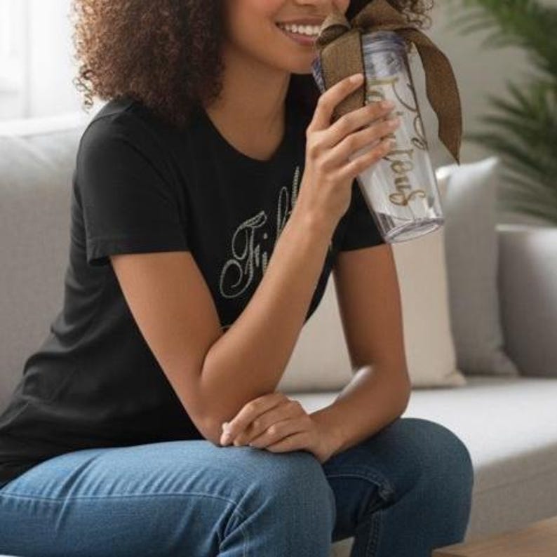 May include: A clear, slender water bottle with gold lettering and a brown ribbon. The bottle is held by a person wearing a black t-shirt with white cursive text and blue jeans. The background includes a gray couch and a green plant.