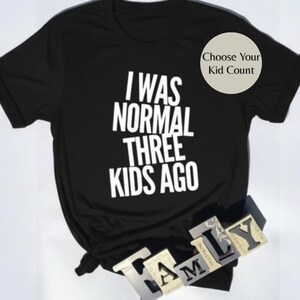 May include: Black t-shirt with the white text "I WAS NORMAL THREE KIDS AGO." A circular graphic in the upper right corner says "Choose Your Kid Count." The word "FAMILY" is spelled out in individual letter blocks at the bottom.