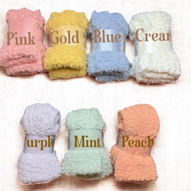 May include: Seven folded pairs of fuzzy socks in pink, gold, blue, cream, purple, mint, and peach. Each pair is secured with a white paper band featuring the corresponding color name printed in brown text across the center of the fabric.