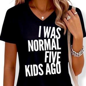 May include: Black V-neck t-shirt with the white text "I WAS NORMAL FIVE KIDS AGO". The shirt is a short-sleeved top, suitable for casual wear. The text is in a bold, sans-serif font.