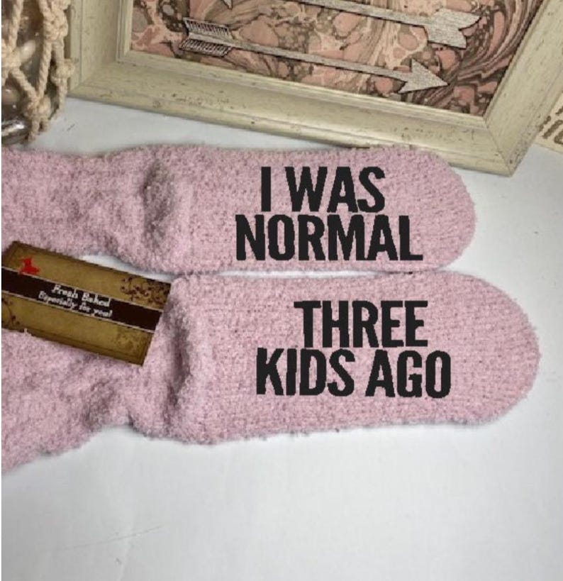 May include: A pair of light pink fuzzy socks with the text "I WAS NORMAL THREE KIDS AGO" in black. A small tag is attached to one sock.