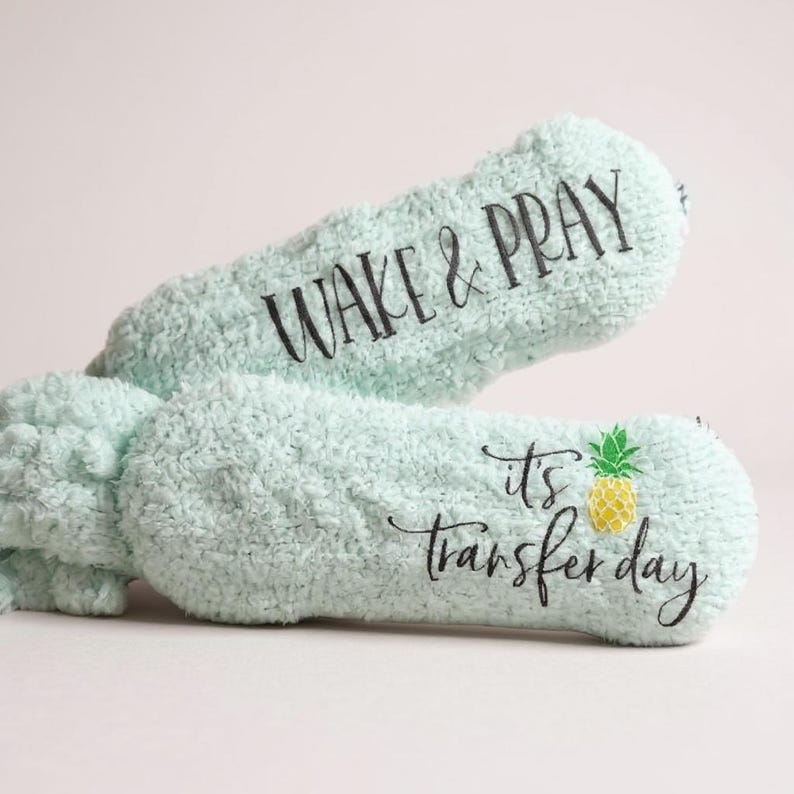 May include: Two light blue, plush eye masks. One mask has the text "WAKE & PRAY" in black script. The other mask has the text "it's transfer day" with a yellow and green pineapple graphic.