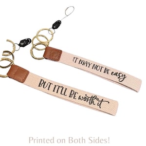 May include: Two keychains with canvas straps and gold-tone rings. The straps are beige with brown leather accents and black text that reads "IT MAY NOT BE easy" and "BUT IT'LL BE worth it".