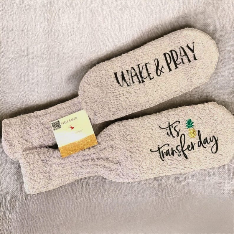 May include: Two light gray, fuzzy slipper socks with the phrases "WAKE & PRAY" and "It's Transfer Day" printed in black script. A small tag with the words "FRESH BAKED" is attached to one sock. The socks are on a light gray surface.