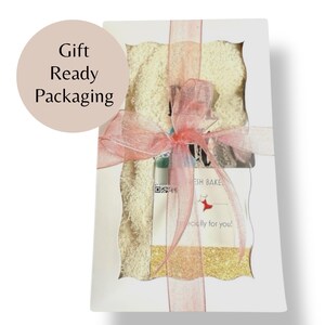 May include: A gift box with a white exterior, tied with a sheer pink ribbon. The box contains a product with a light beige color and gold glitter. The text "Gift Ready Packaging" is in a circle on the left side of the box.