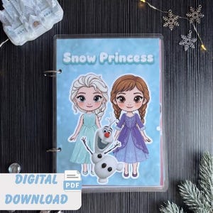 May include: A binder with a blue cover featuring the text "Snow Princess" and illustrations of two cartoon princesses and Olaf the snowman. The words "DIGITAL DOWNLOAD PDF" are at the bottom. The binder is on a dark surface with decorative elements.