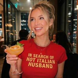 May include: A woman in a red t-shirt with the text "IN SEARCH OF HOT ITALIAN HUSBAND" in yellow. She holds a cocktail with an orange slice and mint garnish. The background shows a restaurant interior.