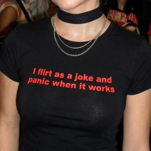 Könnte beinhalten: Schwarzes T-Shirt mit rotem Text: "I flirt as a joke and panic when it works." Die Person trägt einen schwarzen Choker und eine silberne Halskette. Das Bild ist eine Nahaufnahme des T-Shirts und der Accessoires.