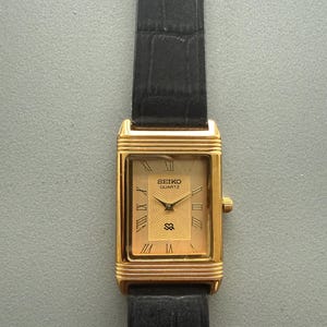May include: A gold-toned Seiko Quartz watch with a rectangular face and black leather strap. The watch face features Roman numerals and the Seiko Quartz logo. The watch has a gold-coloured crown and a textured, black leather strap.