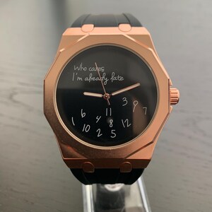 May include: A rose gold-toned wristwatch with a black silicone strap and a black dial. The dial features the text 'Who cares I'm already late' in white script, with scattered white numbers instead of traditional hour markers and rose gold hands.