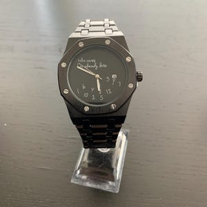May include: A black wristwatch with a metallic link band and an octagonal bezel featuring exposed screws. The black dial displays the text 'Who cares I'm already late' and scattered white numerals, with a date window at the three o'clock position.