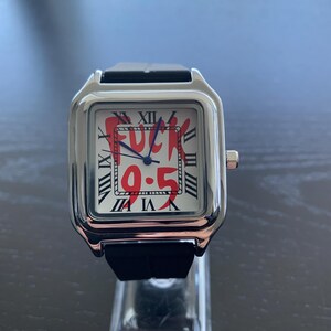 May include: A square wristwatch with a silver-tone metal case and a black silicone strap. The white dial features black Roman numerals around the perimeter, blue hands, and the red text "FUCK 9-5" printed prominently across the centre of the face.