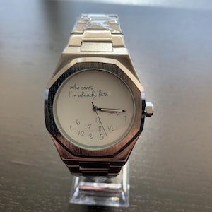May include: A silver-toned stainless steel wristwatch with a white dial. The dial features the text 'Who cares I'm already late' in black script, with scattered, non-sequential numbers. The watch has a metal link band and a protective plastic film.