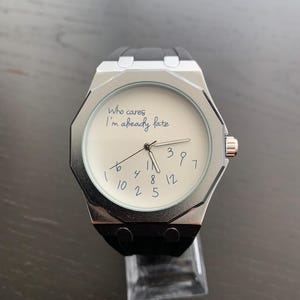 May include: A silver-toned wristwatch with a black rubber strap and an octagonal bezel. The white watch face features the text 'Who cares I'm already late' in blue script, with blue handwritten-style numbers scattered randomly across the dial.