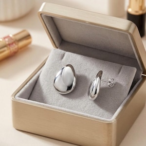 Può includere: Un paio di orecchini a lobo in argento lucido a forma di gocce lisce e arrotondate. Gli orecchini sono esposti all'interno di un portagioie quadrato in metallo color champagne con un rivestimento interno in velluto grigio chiaro e coperchio.