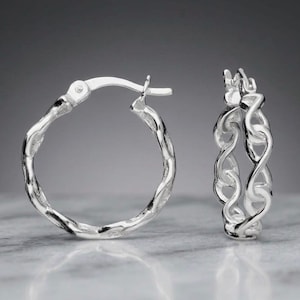 May include: A pair of silver hoop earrings featuring a textured, twisted chain-link design. The earrings have a polished, reflective finish and a hinged post closure mechanism, showcasing a circular shape with an intricate, interconnected metal pattern.
