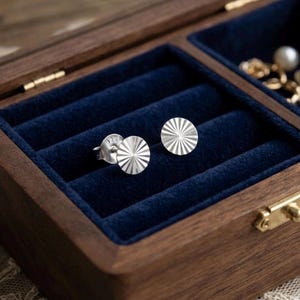 May include: A pair of round sterling silver stud earrings featuring a sunburst pattern of etched lines radiating from the centre. The back of one earring is clearly stamped with the number 925, indicating the silver content of the metal.