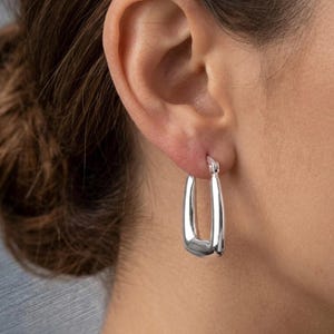 May include: A close-up of a silver-coloured, rectangular hoop earring. The earring is worn on the ear of a person. The earring has a smooth, polished surface and a rectangular shape with rounded corners. The earring is a simple, elegant design.