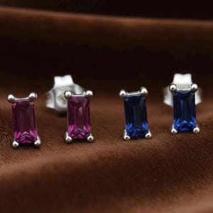 May include: Four stud earrings with rectangular gemstones. Two earrings have magenta-coloured stones, and two have blue stones. The earrings are set in silver-coloured metal on a brown surface.